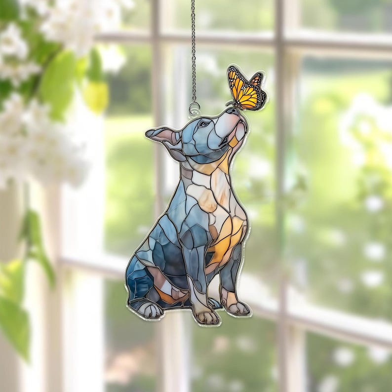 May include: Stained glass dog ornament in shades of blue, gray, and gold, with a butterfly perched on its nose. The dog is sitting and looking upwards. The ornament hangs from a silver chain.