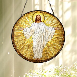 May include: A round stained glass window depicting a figure with outstretched arms, wearing a white robe, and surrounded by a golden halo. The mosaic-style background radiates light. The window is framed in black and suspended by a chain.