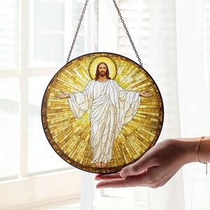 May include: Round stained glass art piece depicting Jesus with outstretched arms, surrounded by a golden halo and background. The artwork features a mosaic-like design with a dark border and is suspended by a chain.