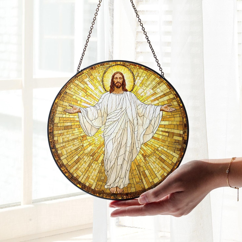 May include: Round stained glass art piece depicting Jesus with outstretched arms, surrounded by a golden halo and background. The artwork features a mosaic-like design with a dark border and is suspended by a chain.
