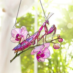 May include: A stained glass suncatcher featuring a pink praying mantis perched on a branch with pink orchids. The insect has a segmented body with purple and pink accents. The suncatcher hangs from a silver chain.