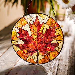 May include: A round stained glass window featuring a vibrant red and orange maple leaf design. The leaf is the focal point, set against a background of yellow and orange geometric shapes. A black chain is attached for hanging.