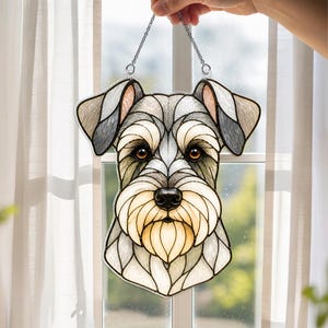 May include: Stained glass Schnauzer dog suncatcher with a silver chain for hanging. The dog's face features shades of gray, tan, and white, with brown eyes and a black nose. The suncatcher is displayed in front of a window with white curtains.