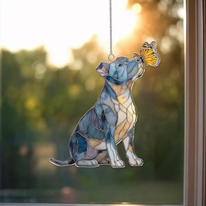 May include: Stained glass dog suncatcher in shades of blue, yellow, and white, with a butterfly perched on its nose. The dog is suspended by a silver chain, creating a decorative window accent. The design has a mosaic appearance.