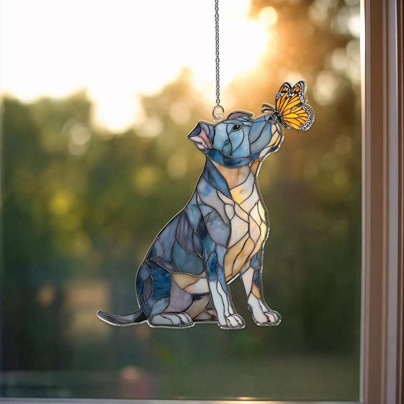 May include: Stained glass dog suncatcher in shades of blue, yellow, and white, with a butterfly perched on its nose. The dog is suspended by a silver chain, creating a decorative window accent. The design has a mosaic appearance.