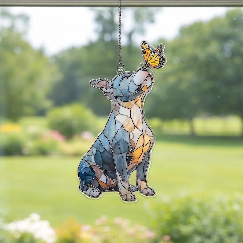 May include: Stained glass window hanging featuring a dog looking up at a butterfly. The dog is primarily blue and tan, with a butterfly in shades of orange and black. The window hanging is suspended by a silver chain, with a green background.