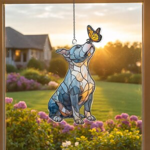 May include: Stained glass window hanging featuring a dog looking up at a butterfly. The dog is blue, gray, and tan, with a butterfly in shades of orange and black. The scene is set against a backdrop of a house and a garden.