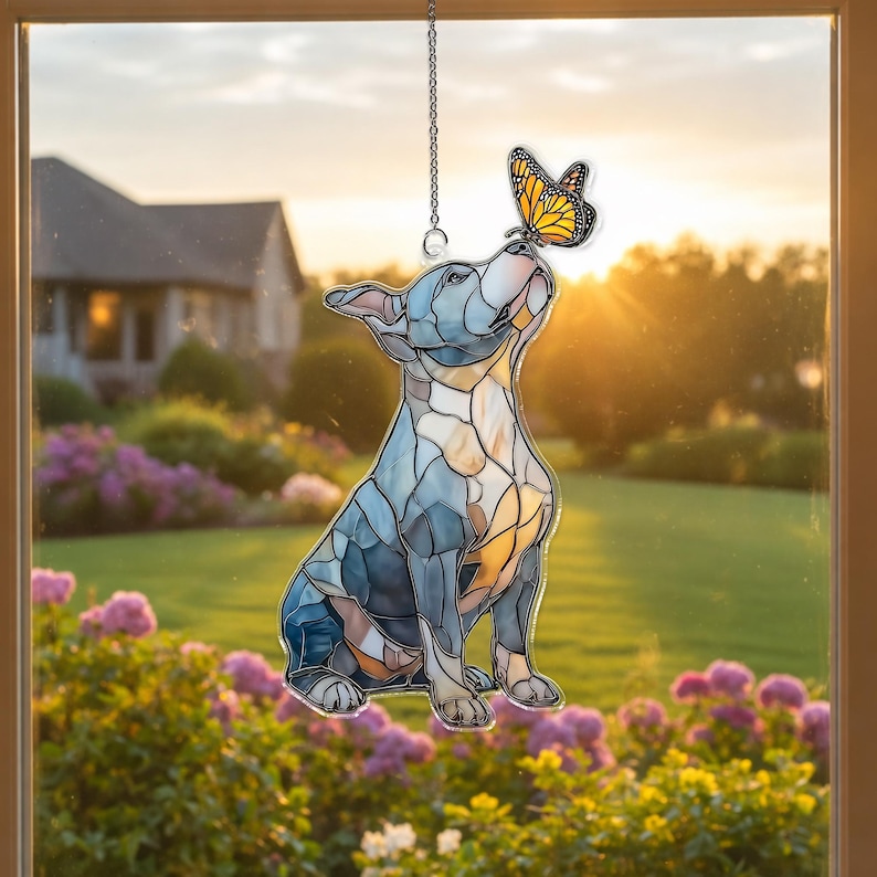 May include: Stained glass window hanging featuring a dog looking up at a butterfly. The dog is blue, gray, and tan, with a butterfly in shades of orange and black. The scene is set against a backdrop of a house and a garden.