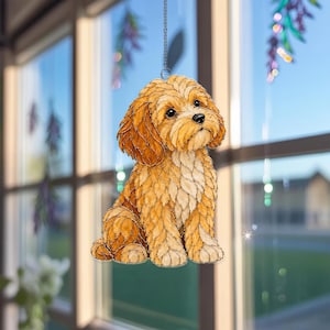 May include: A stained glass suncatcher featuring a golden-brown dog with curly fur, dark eyes, and a black nose. The dog is depicted in a sitting position with its head tilted slightly upward, suspended by a thin silver chain against a window.