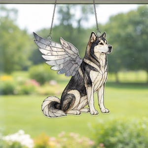 May include: A stained glass suncatcher depicting a sitting husky dog with large, detailed wings. The dog features black, white, and grey glass segments with dark lead lines. The piece is suspended by a thin metal chain for hanging in a window.