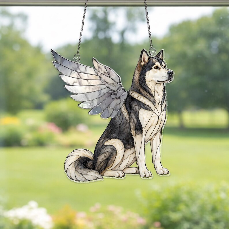 May include: A stained glass suncatcher depicting a sitting husky dog with large, detailed wings. The dog features black, white, and grey glass segments with dark lead lines. The piece is suspended by a thin metal chain for hanging in a window.