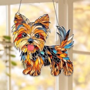 Op de afbeelding: Een glas-in-lood Yorkshire Terrier hond zonnevanger. De hond is afgebeeld in een moza&iuml;ekstijl met tinten oranje, geel, bruin, blauw en zwart. Hij heeft een roze tong en hangt aan een ketting.