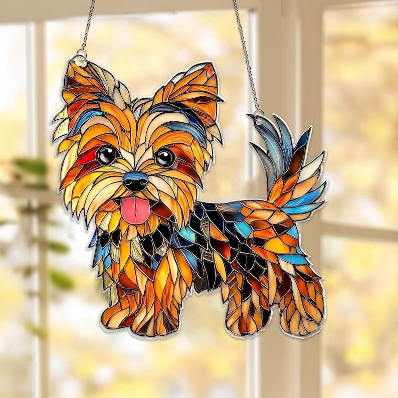 Op de afbeelding: Een glas-in-lood Yorkshire Terrier hond zonnevanger. De hond is afgebeeld in een moza&iuml;ekstijl met tinten oranje, geel, bruin, blauw en zwart. Hij heeft een roze tong en hangt aan een ketting.