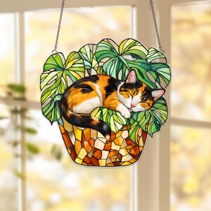May include: A stained glass cat suncatcher. The cat is orange, black, and white, sleeping peacefully on green leaves in a mosaic pot. The suncatcher hangs from a silver chain, perfect for a window or wall.