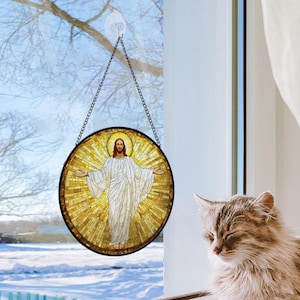 May include: A round stained glass window hanging from a chain, depicting Jesus with outstretched arms. The artwork features a mosaic design with a golden halo and white robes. The window is set against a snowy outdoor scene.