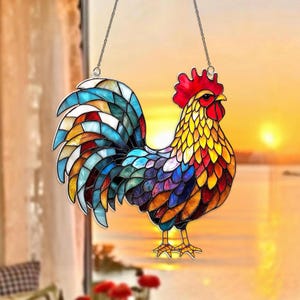 May include: A stained glass rooster suncatcher with a vibrant mosaic design. The rooster features a red comb, yellow and orange body, and a blue, green, and purple tail. It hangs from a silver chain, perfect for window decor.