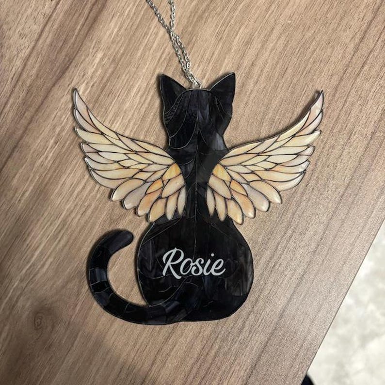 Pu&ograve; includere: Un ornamento a forma di gatto in vetro colorato nero con ali color crema e una coda arricciata, sospeso da una catena argentata. Il gatto presenta il nome Rosie in corsivo bianco sulla schiena, composto da vari segmenti di vetro.