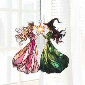 May include: Stained glass suncatcher featuring a princess and a witch holding a star. The princess wears a pink dress and a crown, while the witch is in a green dress and a pointed hat. The suncatcher hangs from a chain.