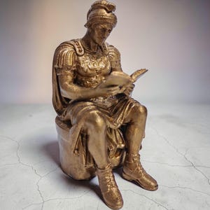 May include: A gold-colored statue of a Roman figure reading a book while seated. The figure wears a helmet, armor, and boots. The statue is sitting on a cylindrical base.