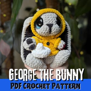 May include: A crocheted stuffed bunny toy with light grey fur, wearing a bright yellow hooded jacket with a single black button. The bunny has large black eyes, a black stitched nose, and holds a small white object. Text reads GEORGE THE BUNNY.