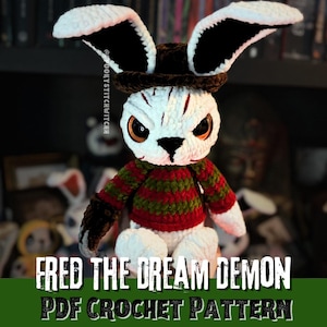 Op de afbeelding: Gehaakte konijnenpop met een wit gezicht, grote zwarte en witte oren en een bruine hoed. De pop draagt een rood-groen gestreepte trui. De tekst op de afbeelding luidt "FRED THE DREAM DEMON PDF CROCHET PATTERN."