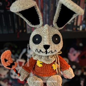 May include: A handcrafted crochet doll, featuring a cream-coloured head and large ears with black interiors. The doll is dressed in an orange sweater with yellow patches and holds a pumpkin-shaped lollipop. Black button eyes and a stitched mouth complete the design.
