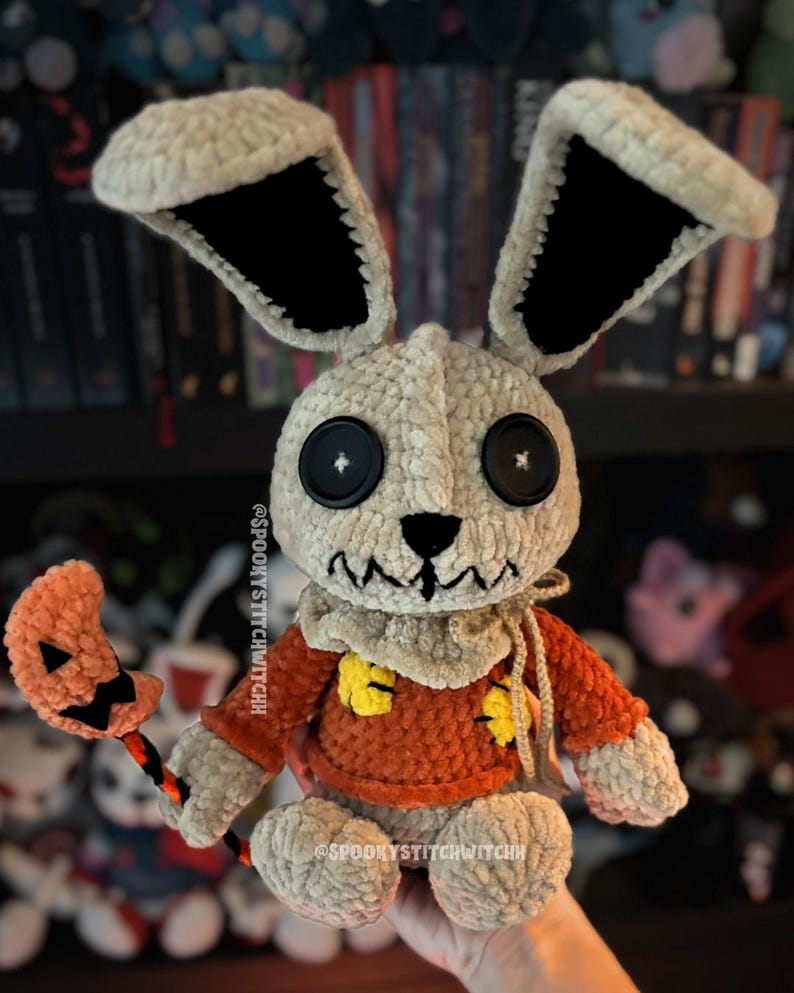 May include: A handcrafted crochet doll, featuring a cream-coloured head and large ears with black interiors. The doll is dressed in an orange sweater with yellow patches and holds a pumpkin-shaped lollipop. Black button eyes and a stitched mouth complete the design.