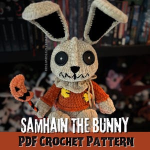 Op de afbeelding: Gehaakte konijnenpop met een schedelachtig gezicht, knoopogen en een gestikte mond. Het konijn draagt een oranje trui en houdt een pompoenvormig accessoire vast. De tekst "SAMHAIN THE BUNNY PDF CROCHET PATTERN" wordt weergegeven.