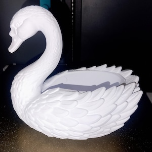 Könnte beinhalten: Ein weißer, schwanförmiger dekorativer Pflanztopf. Der Schwan hat einen langen, gebogenen Hals und detaillierte Federstrukturen. Der Pflanzbereich ist eine flache Schalenform, ideal für kleine Pflanzen oder Dekorationsartikel. Der Schwan besteht aus einem festen, undurchsichtigen weißen Material.