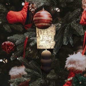 Könnte beinhalten: Eine schwarze Keramik-Tischlampe mit einem weißen Lampenschirm mit Manga-Print. Der Lampenschirm ist rund und mit Schwarzweiß-Illustrationen von Figuren verziert. Die Lampe steht auf einem Weihnachtsbaum mit roten und grünen Ornamenten.