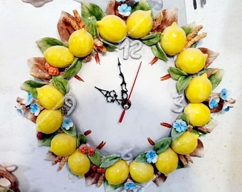 May include: A white ceramic clock with a lemon and leaf design. The clock face has black hands and Roman numeral markings. The clock is surrounded by a wreath of lemons, leaves, and small blue flowers.