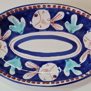Könnte beinhalten: Eine ovale Servierplatte aus Keramik mit blauem Hintergrund und weißem Rand. Die Platte ist mit vier weißen Fischen mit roten und rosa Details sowie grünem Seetang verziert. Die Platte hat einen braunen Punkt-Rand.