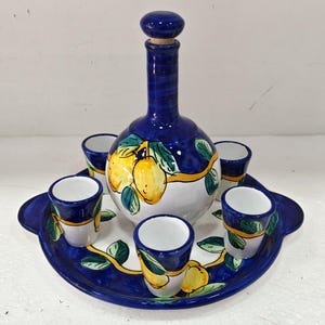 Puede incluir: Un juego de decantador de cerámica que incluye una botella redonda con tapón de corcho y cuatro tazas pequeñas sobre una bandeja ovalada a juego. El conjunto está decorado con limones amarillos, hojas verdes y bordes azul oscuro pintados.