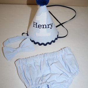 May include: A blue and white striped baby outfit with a matching bow tie and a blue and white striped party hat with the number 1 and the name Henry embroidered on it.