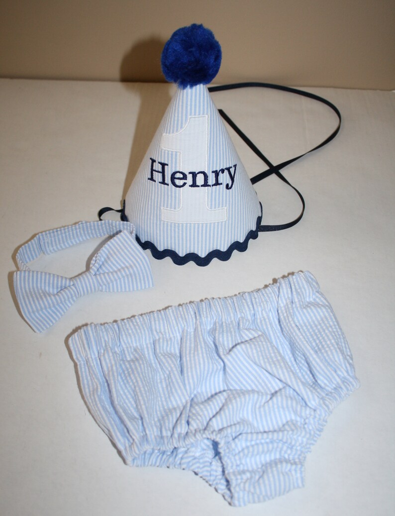 May include: A blue and white striped baby outfit with a matching bow tie and a blue and white striped party hat with the number 1 and the name Henry embroidered on it.