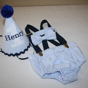 May include: A blue and white striped baby outfit with a matching bow tie and a blue and white striped party hat with the number one and the name Henry embroidered on it.