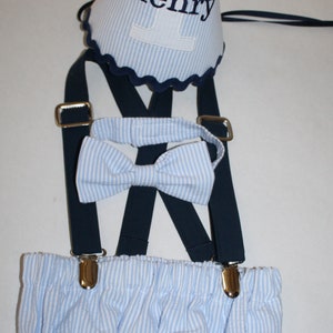 May include: A blue and white striped baby outfit with suspenders, a bow tie, and a blue and white striped party hat with the name "Henry" embroidered on it.