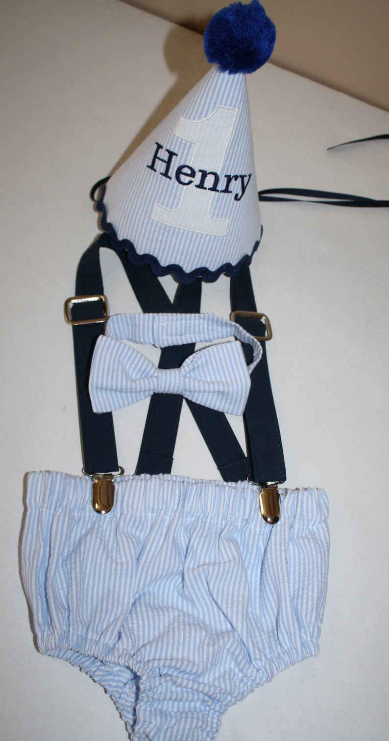 May include: A blue and white striped baby outfit with suspenders, a bow tie, and a blue and white striped party hat with the name "Henry" embroidered on it.