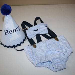 May include: A blue and white striped baby outfit with a matching bow tie and a blue and white striped hat with the name "Henry" embroidered on it.