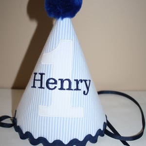 May include: Blue and white striped party hat with a blue pom pom on top. The hat has a white number one and the name "Henry" embroidered on it.
