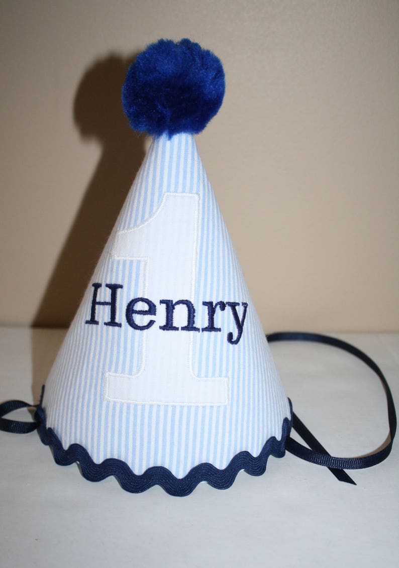 May include: Blue and white striped party hat with a blue pom pom on top. The hat has a white number one and the name "Henry" embroidered on it.