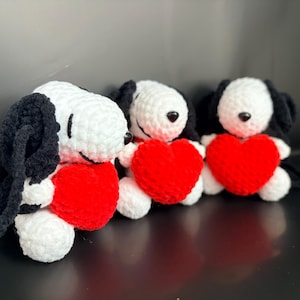 Könnte beinhalten: Drei handgefertigte Snoopy-Plüschtiere aus Häkelarbeit, die jeweils ein rotes Herz halten. Die Plüschtiere haben weiße Körper, schwarze Ohren und schwarze Augen. Der Hintergrund ist dunkel.
