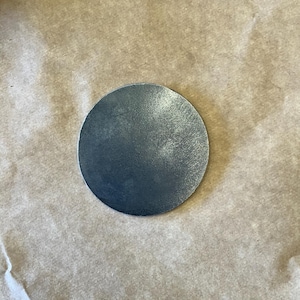 May include: A round, metallic gray object with a textured surface. The object is circular and appears to be made of metal. The background is a light brown, textured surface.