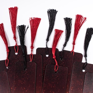 Puede incluir: Un conjunto de diez marcapáginas con borlas rojas y negras. Los marcapáginas están hechos de cartulina marrón oscuro con un sutil efecto de brillo.