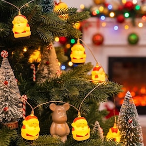 Pode incluir: Um cordão de luzes de Natal com pequenas figuras iluminadas de Pai Natal com gorros vermelhos e rostos amarelos. As luzes estão ligadas por um fio fino e transparente e estão dispostas nos ramos de um pinheiro verde.