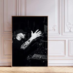 Puede incluir: Fotografía en blanco y negro de un DJ actuando en un concierto. El DJ lleva una gorra negra y una camisa negra con tatuajes en el brazo. Está de pie detrás de una mesa de mezclas y está haciendo un gesto con la mano.