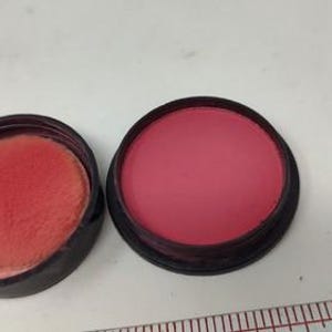 May include: Open blush compact with a dark rim, revealing a vibrant pink powder. The compact is next to a matching applicator pad. A small, round, turquoise-colored object is visible to the left. A ruler is in the bottom right corner.