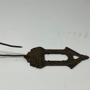 May include: A rusty metal hook with an ornate, arrow-shaped design. The hook extends from a decorative plate with a detailed, textured surface. The metal has a weathered appearance, suggesting age and use. The overall design is symmetrical.