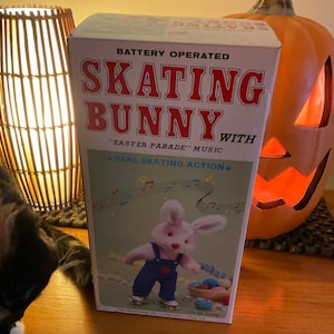 May include: A white box with a red and blue illustration of a skating bunny. The box reads "Battery Operated Skating Bunny with "Easter Parade" Music *Real Skating Action*"