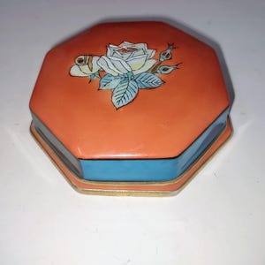 May include: An octagonal trinket box with a coral-colored lid, featuring a white rose with blue-green leaves. The box has a light blue band and gold trim, creating a vintage aesthetic. A decorative storage item.